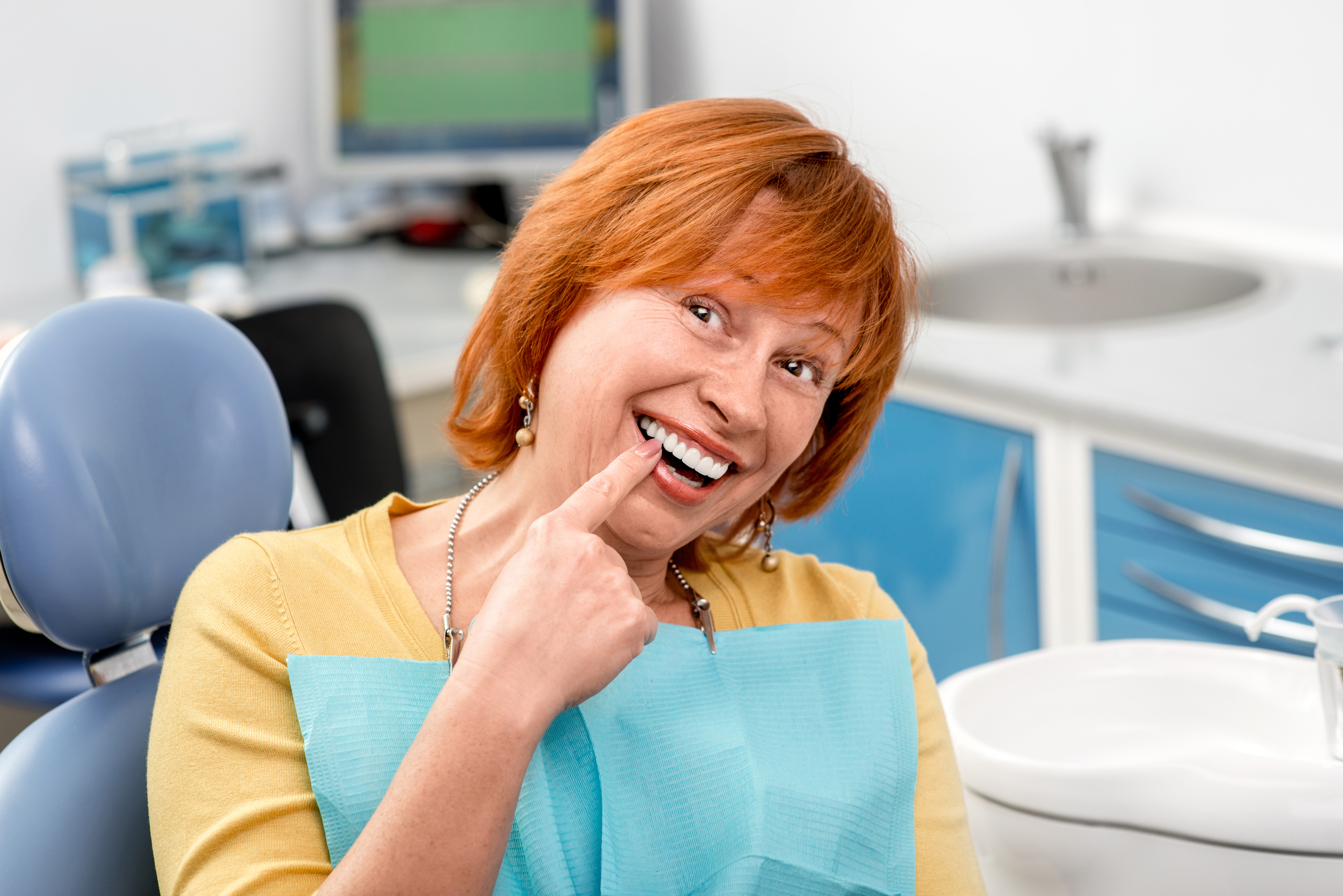 The image shows a woman with red hair, wearing a yellow shirt, sitting in a dental chair with her mouth open as if she's speaking or singing. She is holding a blue object that could be a toothbrush or a dental tool near her mouth. Behind her, there is a dental office environment with a sink and other dental equipment visible.