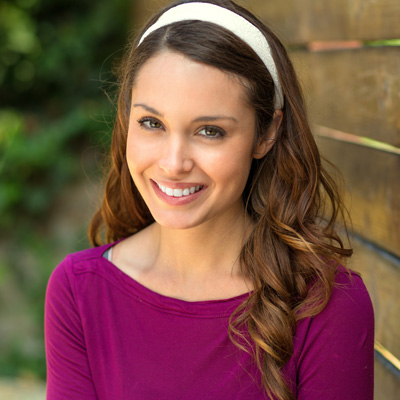 The image is a portrait of a woman with long hair, wearing a purple top and a headband. She is smiling at the camera and posing against a wooden fence background.