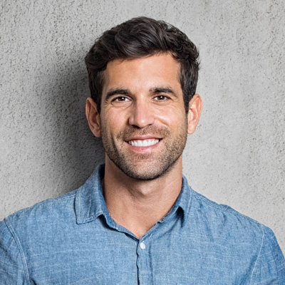 A smiling man with short hair, wearing a blue denim shirt and standing against a gray wall.