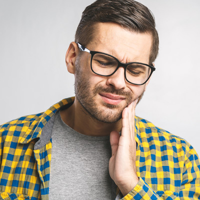 The image features a man with glasses, sporting a beard and mustache, wearing a plaid shirt and holding his hand to his chin. He appears to be in a state of distress or concern, possibly due to dental pain.