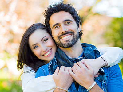 The image is a photograph of two adults, possibly a couple, smiling and posing together. They appear to be in a happy mood, with the man wearing a short beard and the woman having curly hair. Both are dressed casually, with the man in a T-shirt and the woman in a top with lace sleeves. The background is plain, suggesting that the photo might have been taken indoors or at an event.