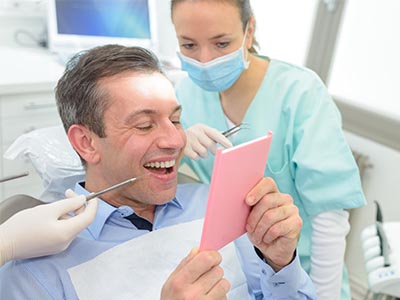 A man is seated in a dental chair, holding a pink card and smiling broadly while wearing glasses. Behind him, a female dentist is attentively looking at the card with a slight smile on her face.