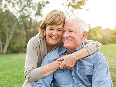 An elderly couple, a man and a woman, are embracing each other in a park setting.