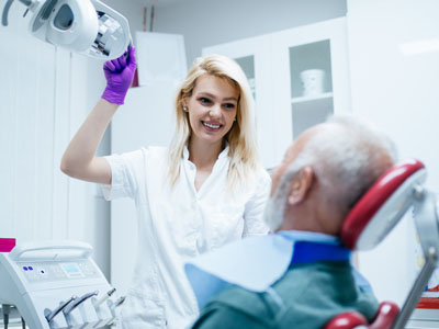 A woman in a white coat stands by an elderly man receiving dental care, holding a device and smiling.