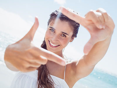 The image features a young woman with long hair, smiling and holding up her hand in front of her face, creating a frame with her fingers. She is outdoors, wearing a white top, and appears to be enjoying a sunny day at the beach.